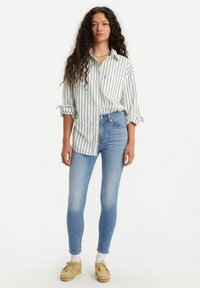 Camisa blanca de manga larga con botones y rayas verticales negras, combinada con jeans ajustados azules y zapatos beige, con las mangas arremangadas.