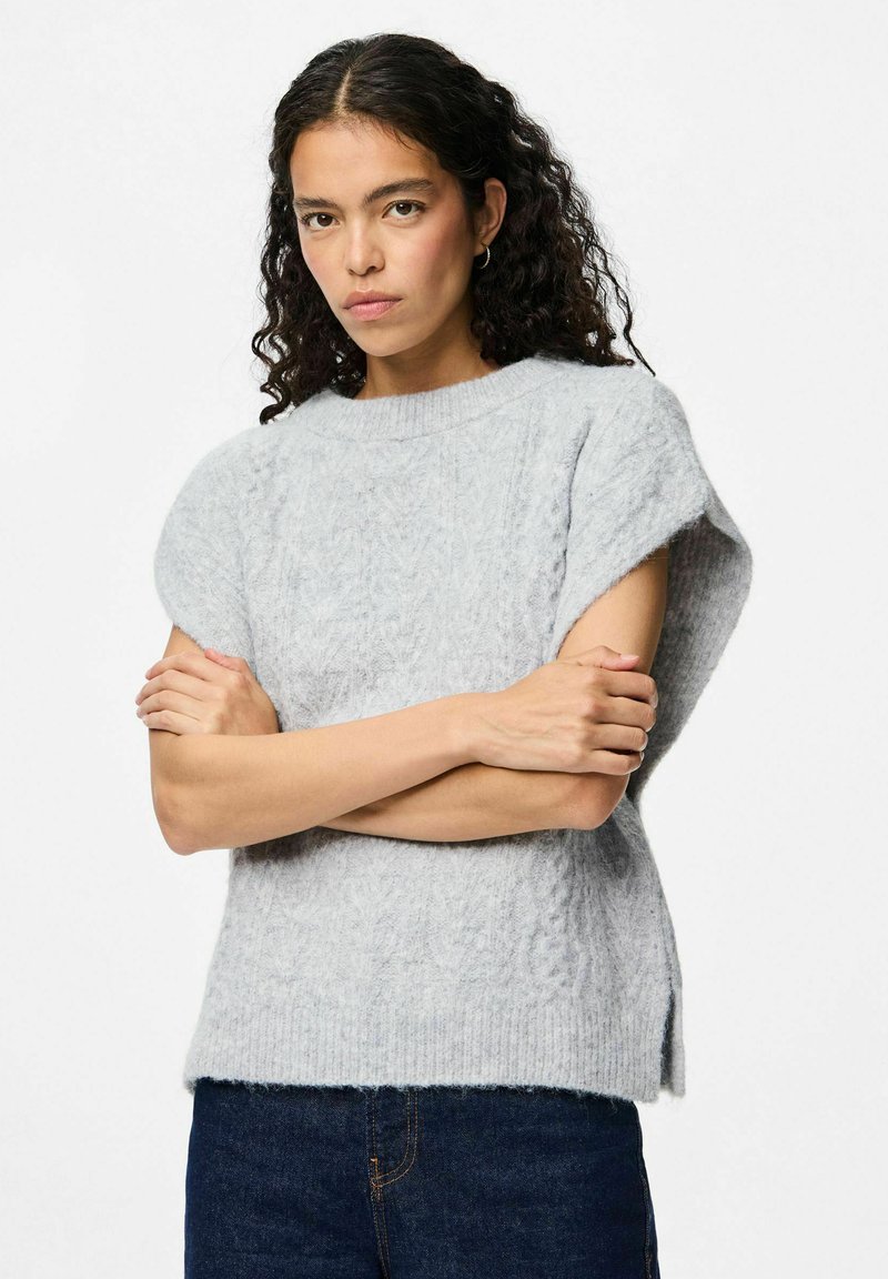 Ljusgrå kabelstickad tröja med korta ärmar, ribbad halsringning och sidofack. Mjuk textur, avslappnad passform, matchad med mörka jeans.