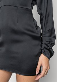 Mustan satiinimekko, jossa on pitkät, kerätty hihat ja muotoiltu vartalo-osa. Ominaisuuksiin kuuluu suora helma ja sivutaskut. Sujuva pinta.