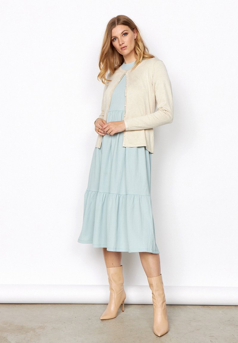 Beige kardigan s knoflíky přes světle modrou, texturovanou, vrstvenou šaty. Outfitu dominují světle béžové jehlové boty.