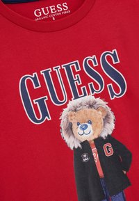 Crvena pamučna majica s crtić medvjedom s lavljom grivom u crnom kaputu, odvažno "GUESS" logo u mornarsko plavoj boji iznad grafike.