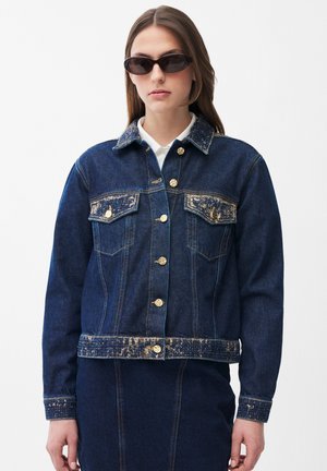 Donna che indossa una giacca in denim blu scuro con bottoni color oro e dettagli consumati, abbinata a una gonna in denim coordinata e occhiali da sole neri.