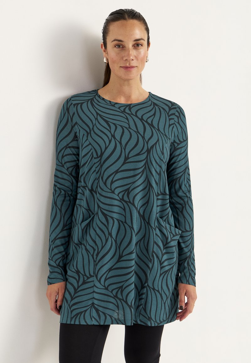 Teal lange mouw tuniek met een zwart golfpatroon, ronde halslijn en zijzakken. Zacht, stretchoverend materiaal; relaxte pasvorm voor comfort.