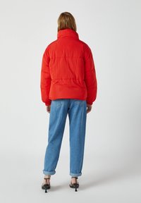 Rød puffjakke med høj krave, quilted struktur og elastiske manchetter; parret med opsmøgede lyseblå jeans og sorte hæle-sandaler.