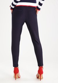 Person in navyblauen Nadelstreifen-Hosen und roten High Heels, mit Händen in den Taschen, von hinten vor einem weißen Hintergrund gezeigt.