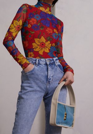 Osoba ubrana w jasnoniebieskie jeansy z wysokim stanem i przezroczysty czerwony top z długim rękawem w kwiaty, trzymająca beżową torebkę z nadrukiem sygnalizacji świetlnej.