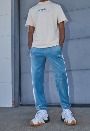Homme portant une chemise beige à manches courtes et un jogging en velours bleu avec des bandes blanches sur les côtés, debout devant une porte de garage grise les mains dans les poches.