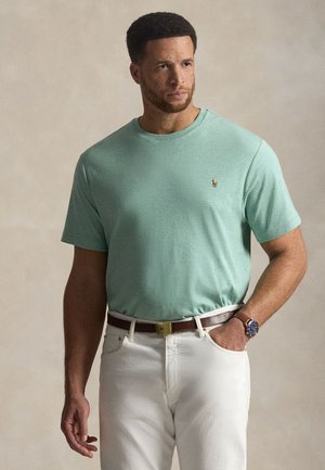 Miętowa koszulka z krótkim rękawem i okrągłym dekoltem, z małym logo na piersi, w połączeniu z białymi dżinsami i brązowym paskiem.