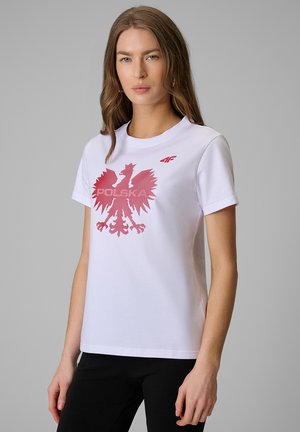 Kobieta z długimi brązowymi włosami nosi białą koszulkę z czerwonym polskim orłem i czarne spodnie na jednolitym szarym tle.