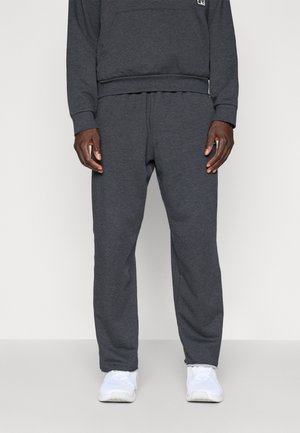 Mann trägt dunkelgraue Jogginghose und passendes Sweatshirt mit weißen Sportschuhen und steht vor einfachem Hintergrund.