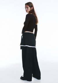 Fekete hosszú ujjú felső, amelyet széles szárú fekete nadrággal viselnek, a nadrág réteges szegéllyel, nyers széllel és derékszíjjal rendelkezik. Simalebújású anyag.