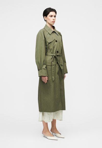 Χακί χακί μπουφάν σε ελαφρύ ύφασμα με ζώνη που δένει, μεγάλα μπροστινά τσέπες και κούμπωμα με κουμπιά. Λευκά επίπεδα παπούτσια και στρωμένο ανοιχτό φόρεμα από κάτω.