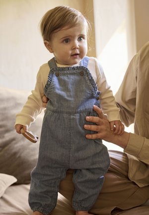 Lichteblauwe denim tuinbroek met verstelbare banden en houten knopen. Gecombineerd met een crèmekleurig shirt met lange mouwen. Zachte textuur, losse pasvorm.