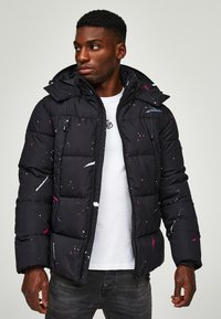 Μαύρο μπουφάν puff με πολύχρωμες σχέσεις πιτσιλιές, διαθέτει κουκούλα, κλείσιμο με φερμουάρ και ελαστικούς μανσέτες. Φοριέται πάνω από ένα λευκό πουκάμισο.