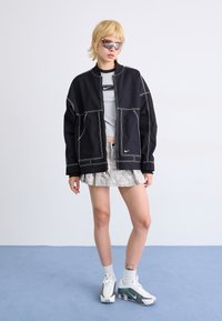 Casaco preto com costura branca, blusa cinza, saia com padrão metálico e ténis cinza e brancos. Modelo usa óculos de sol com cor.