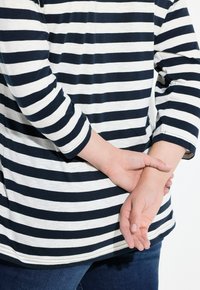 Gestreifte Langarm-Bluse in Marineblau und Weiß, mit lockerer Passform und abgerundetem Saum. Denim-Jeans sind sichtbar, die Hände sind hinten verschränkt.