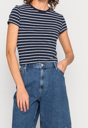 Vrouw draagt een marineblauw-wit gestreept crop top met korte mouwen, ingestopt in een high-waisted blauw spijkerbroek, met één hand in de zak.
