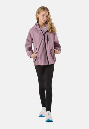 Ragazza giovane con capelli lunghi e ricci, che indossa una giacca con cappuccio color malva, leggings neri e sneakers bianche con suola blu, in piedi con le mani nelle tasche.