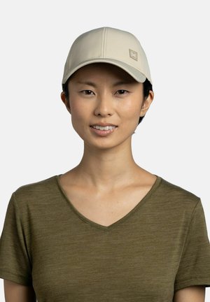 Woman with short black hair wearing a beige cap with a logo and an olive green V-neck T-shirt, smiling against a light background.