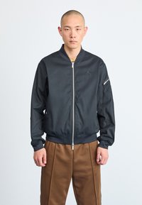 Melna bomber jaka ar rāvējslēdzēju priekšpusē, ar ribot�ām piedurknēm un apakšmalu, ar mazu logotipu uz krūšu daļas un sānu kabatu ar rāvējslēdzēju.
