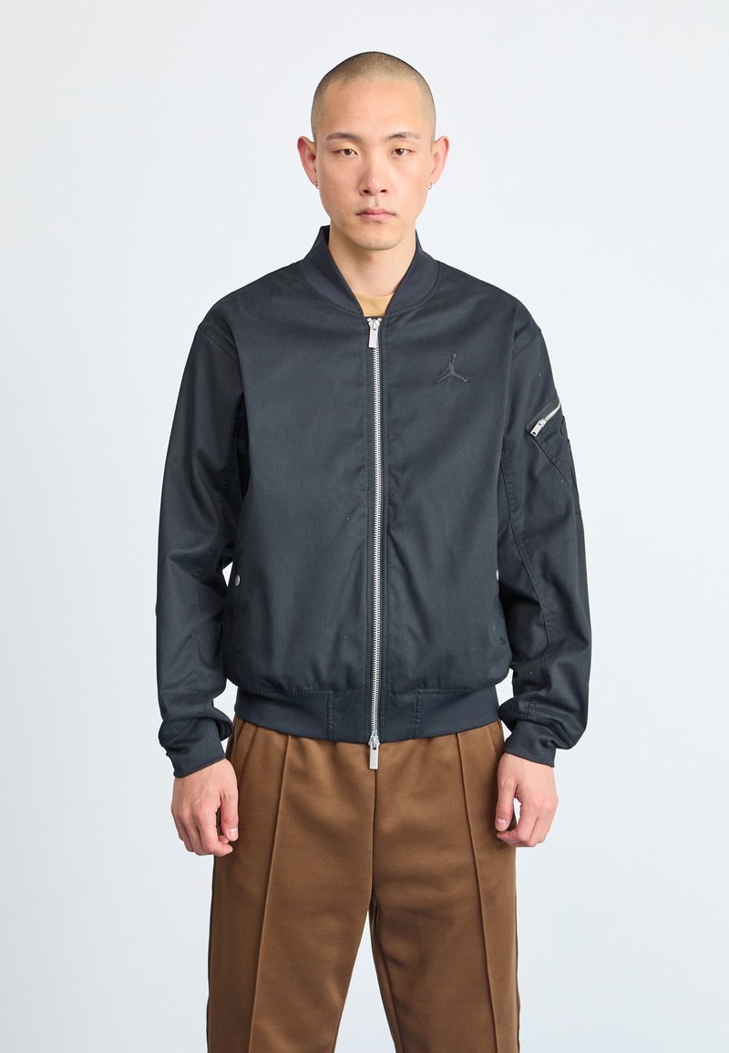 Melna bomber jaka ar rāvējslēdzēju priekšpusē, ar ribotām piedurknēm un apakšmalu, ar mazu logotipu uz krūšu daļas un sānu kabatu ar rāvējslēdzēju.