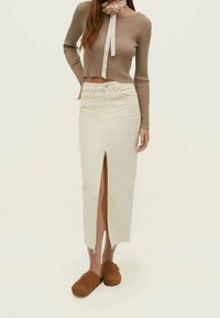 Knee-langer cremefarbener Jeansrock mit vorderem Schlitz und ausfransendem Saum, kombiniert mit einem taillierten, sandfarbigen Rippsweater und braunen Holzclogs.