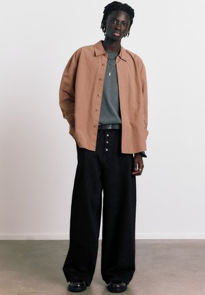 Giovane persona con pelle scura e capelli intrecciati indossa una camicia a quadri marroni sopra una maglietta grigia, pantaloni larghi neri e scarpe nere, in piedi all'interno.