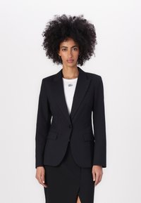 Černý blazer s dvěma předními kapsami, zapínání na jeden knoflík a špičatými klopami. Nositý přes bílé upnuté triko.