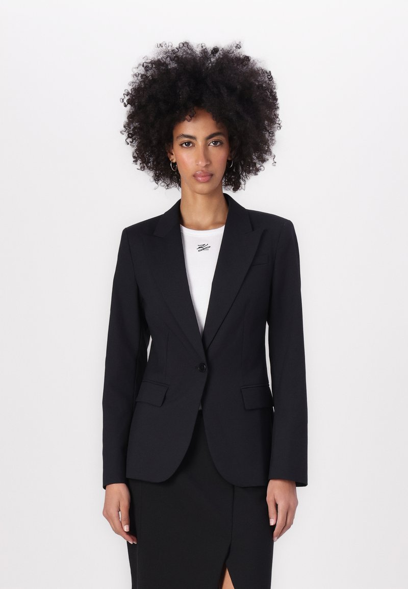 Černý blazer s dvěma předními kapsami, zapínání na jeden knoflík a špičatými klopami. Nositý přes bílé upnuté triko.
