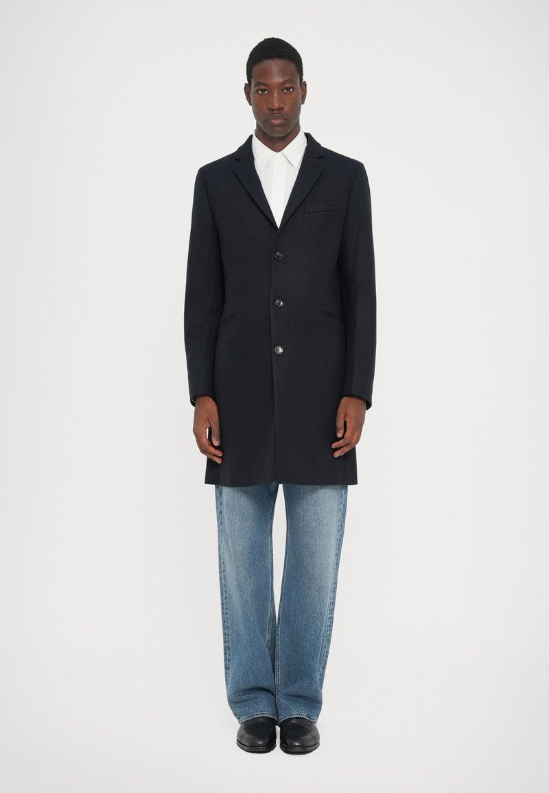 Cappotto in lana blu navy con design a un petto, cinque bottoni, vestibilità sagomata e tasche laterali, abbinato a pantaloni in denim chiaro.