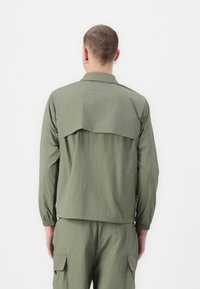 Jachetă verde cu mânecă scurtă, cu o croială lejeră, având un yoke pe spate și manșete elastice, realizată dintr-un material texturat ușor.