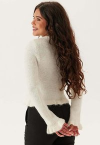 Vit, texturerad tröja med långa ärmar, med fransiga manschetter och en figurnära silhuett, bärs tillsammans med svarta byxor, visat från baksidan.