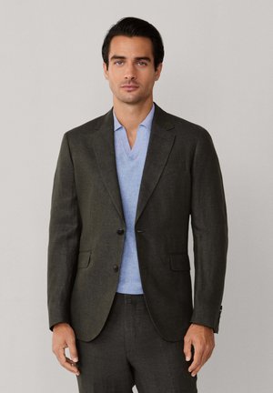 Hombre con chaqueta y pantalones de traje color oliva oscuro, con suéter de cuello en V azul claro, de pie frente a un fondo liso gris claro.