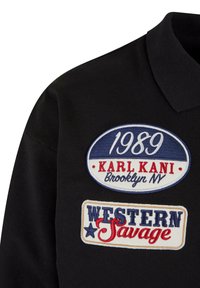Tricou polo negru cu patch-uri brodate. Un patch afișează "1989 KARL KANI Brooklyn NV" în albastru și roșu, iar altul scrie "WESTERN Savage" în albastru și roșu.