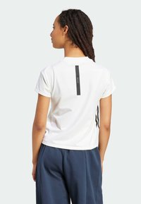 Balts īsām piedurknēm t-krekls ar melnu vertikālu svītru un logotipa detaļu aizmugurē. Gluda auduma, brīva piegriezuma, kombinēts ar ziliem biksēm.