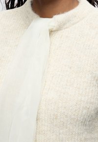 Gros plan d'une personne portant un pull en maille crème avec une écharpe blanche transparente nouée lâchement autour du cou.