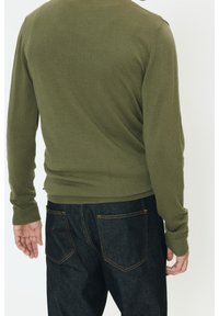 Pull en maille côtelée vert olive à manches longues, coupe ajustée, associé à un jean en denim foncé avec des coutures dorées sur les poches.
