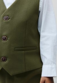 Gilet vert olive en tissu texturé, avec trois boutons marron et une seule poche poitrine, associé à une chemise à manches blanches.