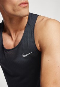 Débardeur de sport Nike en tissu gris foncé, présentant une texture respirante, un design sans manches et un logo réfléchissant sur la poitrine gauche.