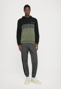 Sweat à capuche noir, gris et vert olive, avec une poche frontale. Associé à des joggers gris avec des rayures noires et olive. Baskets blanches.