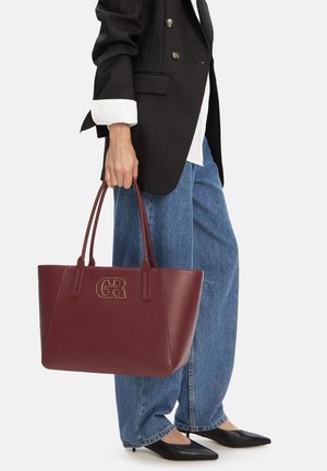 Person trägt ein schwarzes Sakko und blaue Jeans und hält eine große burgunderrote Einkaufstasche mit goldenem "GR"-Logo, kombiniert mit schwarzen spitzen Schuhen.