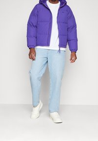 Fialová prošívaná bunda s kapucí a elastickými manžetami; kombinovaná s světle modrými džínami a bílými teniskami.