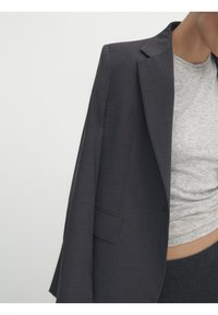 Blazer grigio scuro con vestibilità su misura, sottile texture e rever a punta. Tasca frontale singola e design minimalista. Indossato sopra un top grigio.
