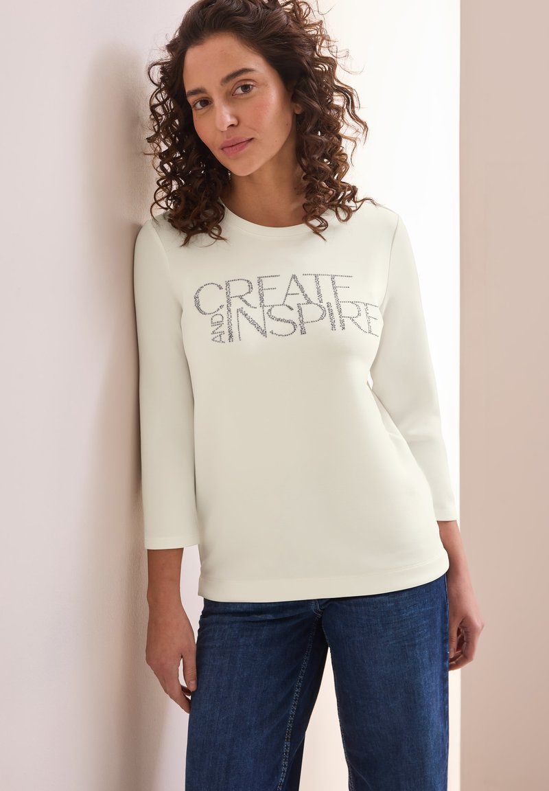 Cream long-sleeve top with "CREATE AND INSPIRE" text in silver embellishment, paired with dark blue jeans. Smooth texture and classic fit.