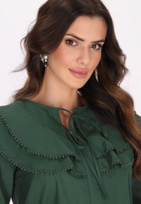 Grüne Bluse mit Rüschendetails und einem Halsband. Glattes Material mit zarten Verzierungen entlang der Rüschen und des Ausschnitts.