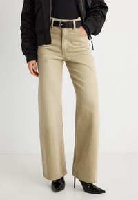 Γυναίκα που φοράει μπεζ τζιν με φαρδιά μπατζάκια, μαύρη ζώνη, μαύρες μυτερές μπότες με ψηλό τακούνι και μαύρο μ πουφάν με το χέρι στην τσέπη.