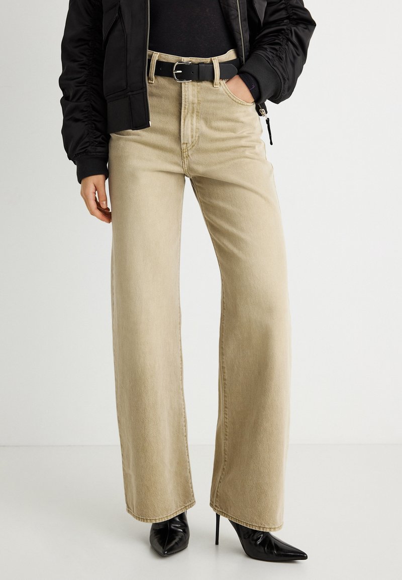 Γυναίκα που φοράει μπεζ τζιν με φαρδιά μπατζάκια, μαύρη ζώνη, μαύρες μυτερές μπότες με ψηλό τακούνι και μαύρο μπουφάν με το χέρι στην τσέπη.