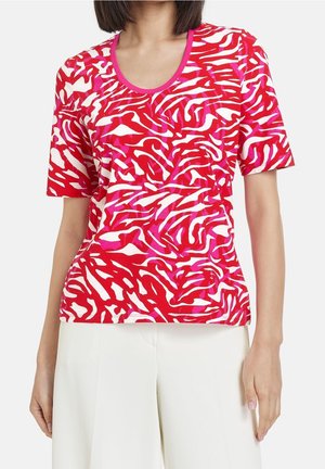 Kobieta ubrana w krótki rękaw, czerwoną, białą i różową bluzkę w abstrakcyjny wzór z różowym dekoltem i białe spodnie.