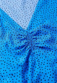 Tissu bleu avec de petits cœurs noirs, présentant un décolleté en V et des fronces au centre. Texture lisse, matériau léger.