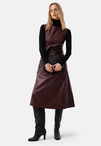 Abito in pelle bordeaux con design asimmetrico, indossato sopra un dolcevita nero, abbinato a una borsa nera intrecciata e stivali al ginocchio neri con texture.